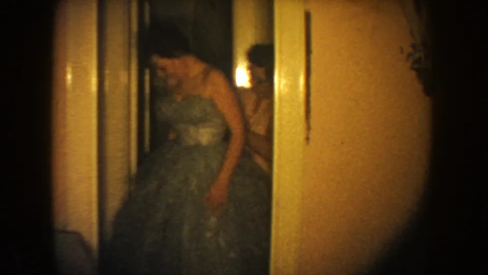 1954 South Dakota 8mm frame showing a woman in a full-skirted gown bending slightly in a private interior hallway.