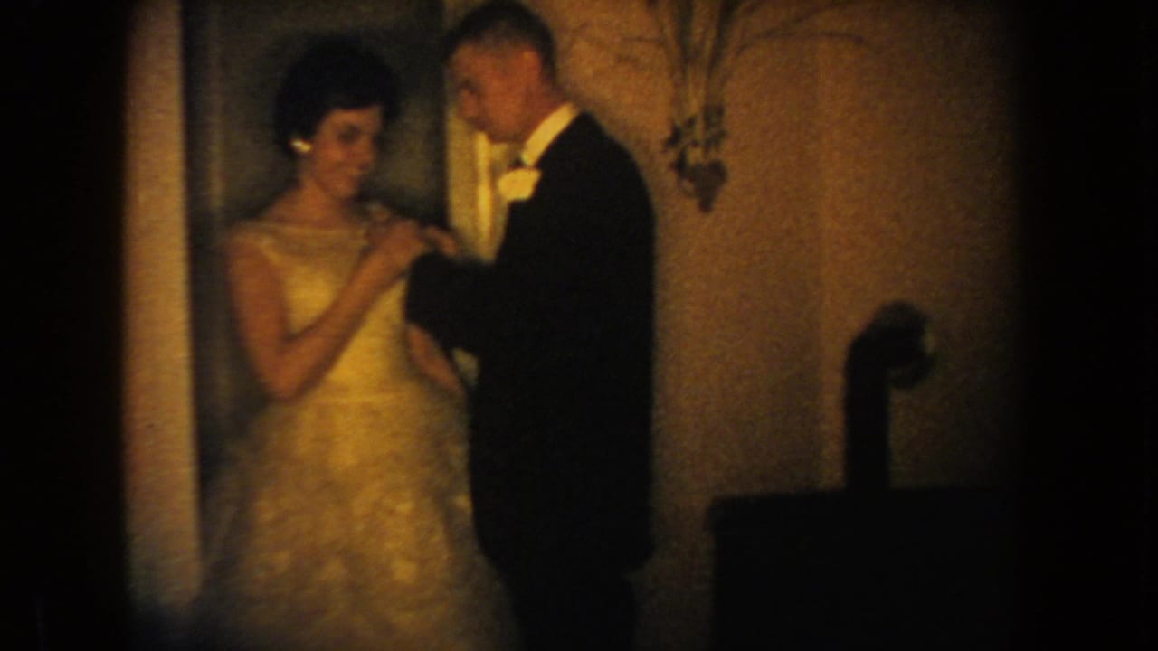 1954 South Dakota 8mm frame showing a woman in a formal gown with a man in a tuxedo and white bow tie indoors.