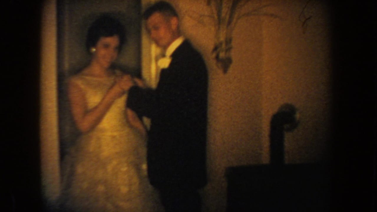 1954 South Dakota home-movie frame showing a tuxedoed man placing a small flower on a woman's light formal gown indoors.