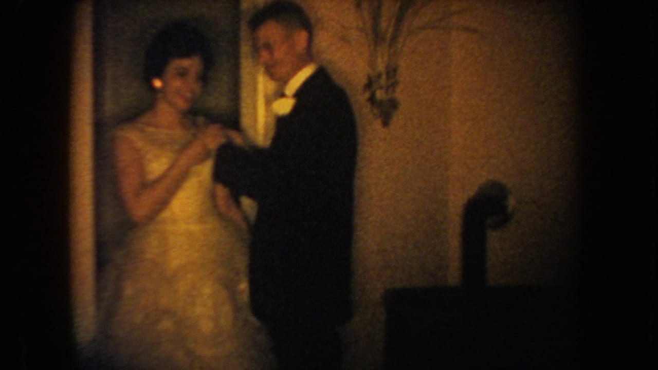 Smiling woman in a light gown stands beside a man in a tuxedo as he works near her bodice.