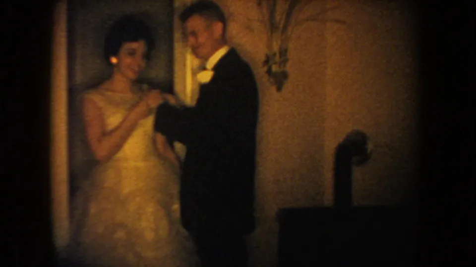 1954 South Dakota 8mm frame of a formal couple indoors, with the man adjusting a flower on the woman's dress.