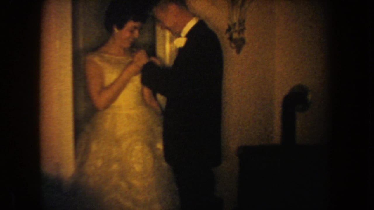 Soft 1954 South Dakota home-movie frame showing a man in formalwear leaning in to fasten a flower to a woman's gown.