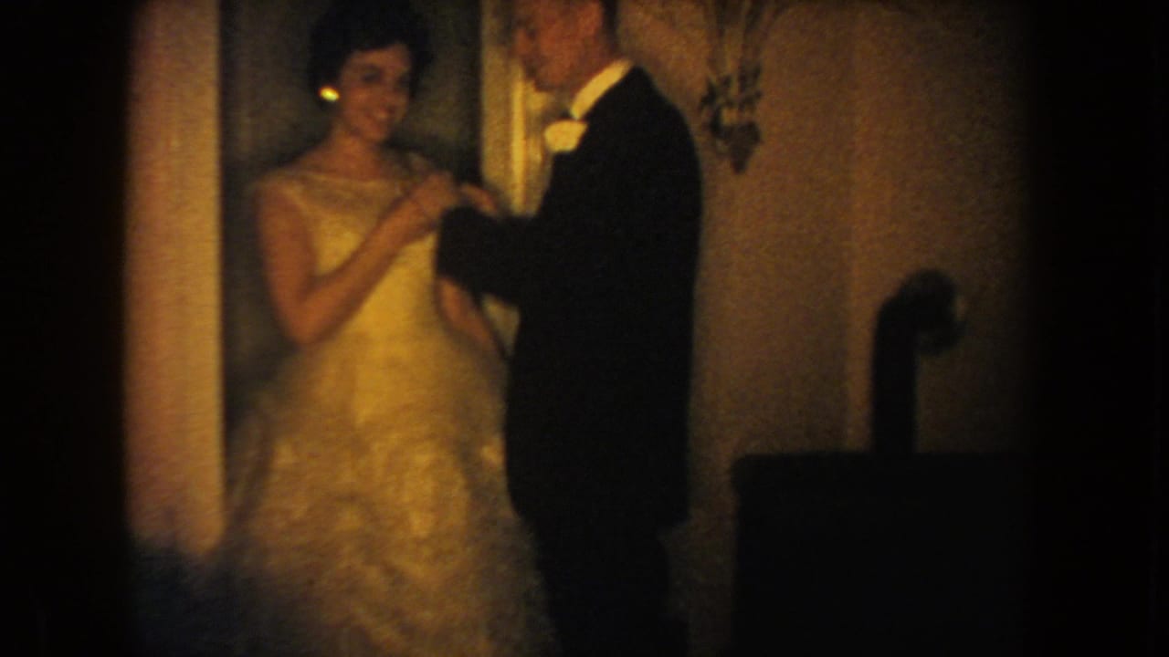 1954 South Dakota frame of a tuxedoed man and woman in a light gown during a quiet flower-adjusting moment indoors.