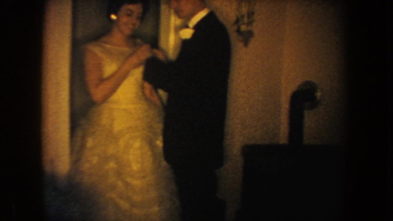 1954 South Dakota home-movie frame of a bride in a white gown and groom in a dark tuxedo standing together indoors.