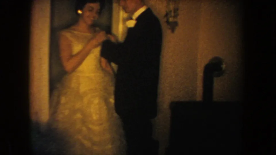 1954 South Dakota home-movie frame of a bride in a white gown and groom in a dark tuxedo standing together indoors.