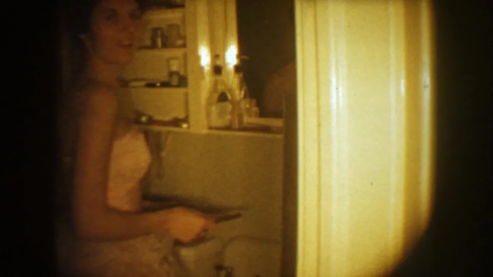 1954 South Dakota home-movie frame of a bride in a bathroom, seen near a sink, mirror, and wall lights.