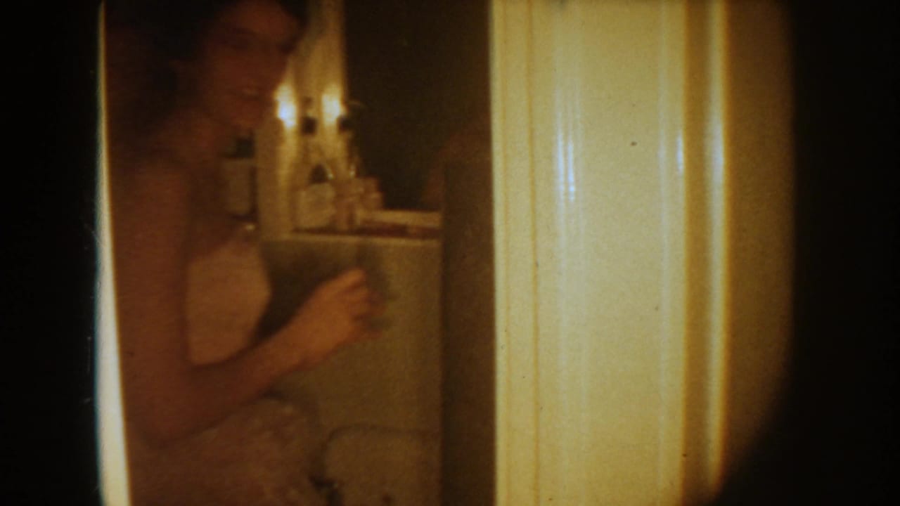 1954 South Dakota home-movie frame showing a woman in a white wedding dress near a bathroom mirror, with dim warm lighting.