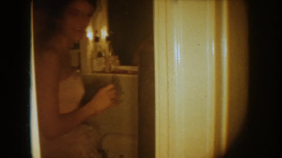 1954 South Dakota home-movie frame showing a woman in a white wedding dress near a bathroom mirror, with dim warm lighting.