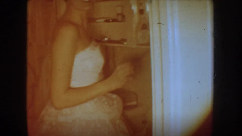 1954 South Dakota home-movie frame showing a closer, soft-focus view of a woman in a white wedding dress inside a bathroom.
