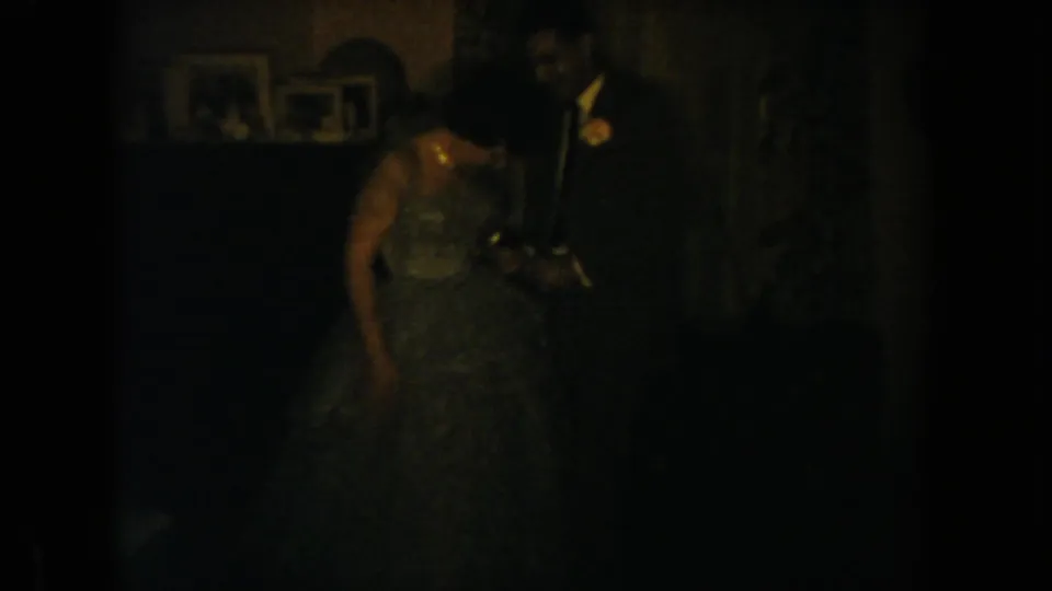 A grainy 1954 South Dakota home-movie frame shows the wedding couple close together in a dark private interior.