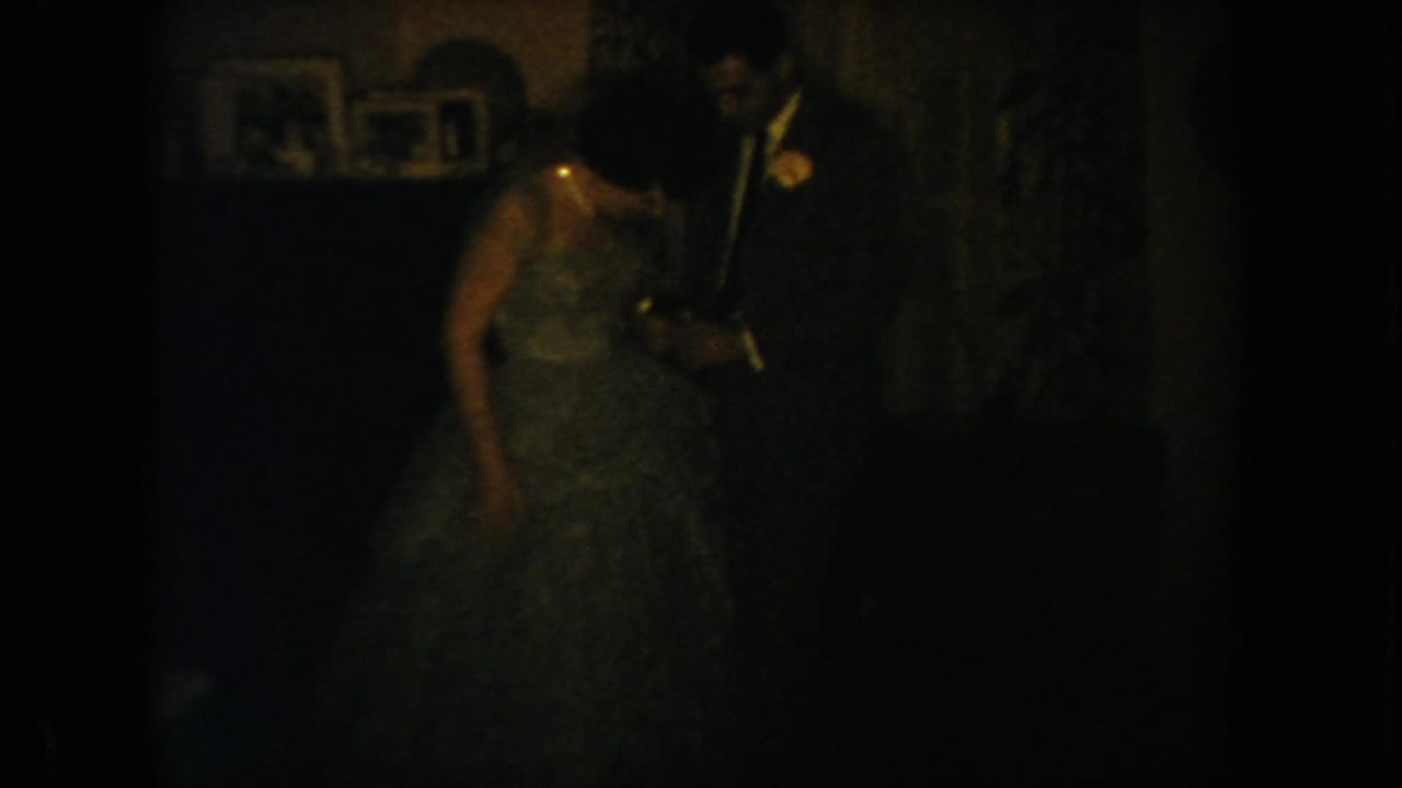 1954 South Dakota home-movie frame showing a couple in formal attire, partly obscured by low light and film grain.