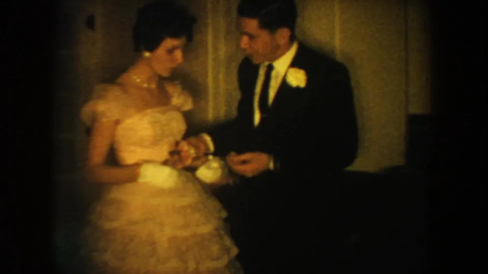 1954 South Dakota home-movie frame of a woman in a ruffled gown and gloves standing beside a man in a dark suit.