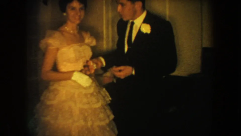 1954 South Dakota home-movie frame showing the couple clearer, with gown, gloves, suit, boutonniere, and dim interior visible.