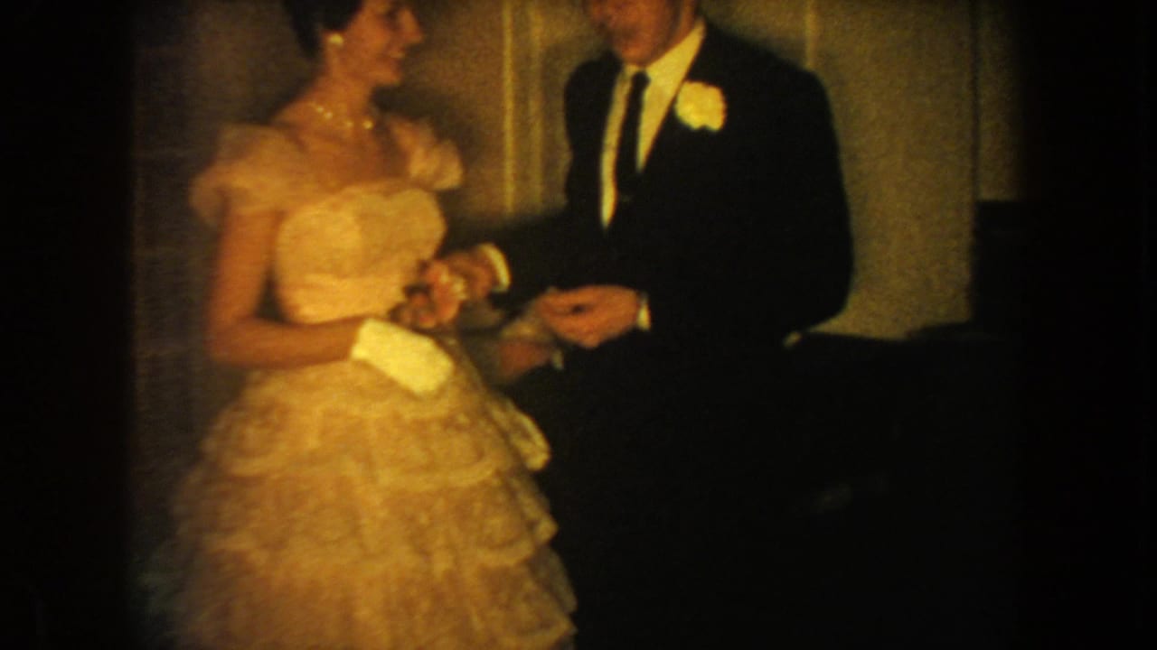In 1954 South Dakota home-movie footage, a formally dressed woman in a ruffled gown stands with a man wearing a dark suit and boutonniere.