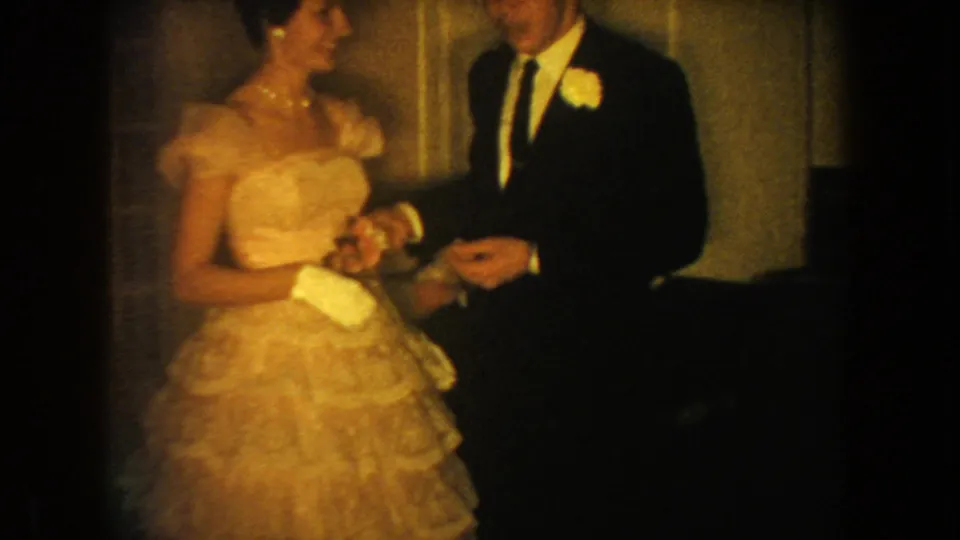 In 1954 South Dakota home-movie footage, a formally dressed woman in a ruffled gown stands with a man wearing a dark suit and boutonniere.