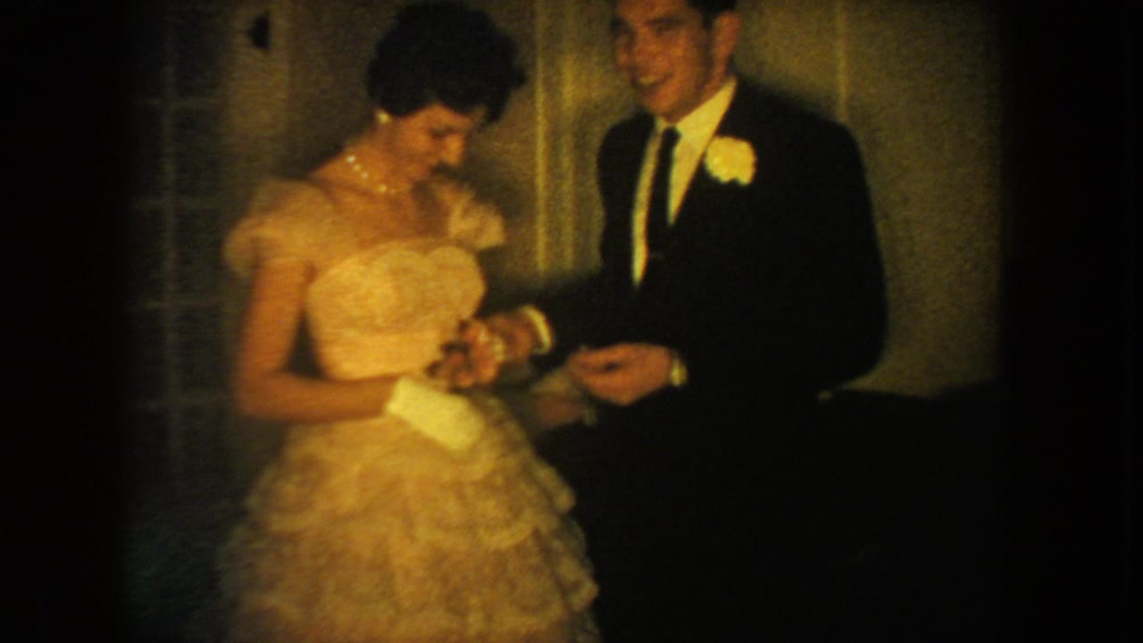 Formal couple smiles while holding hands in a softly lit interior.