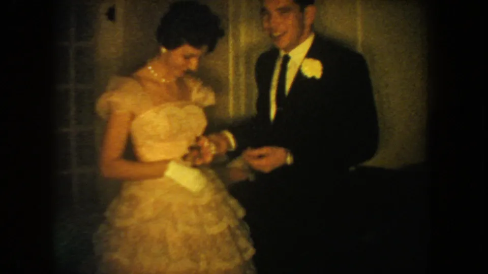 The woman in a ruffled gown looks down while holding hands with a suited man, with warm 8mm grain and soft indoor exposure.
