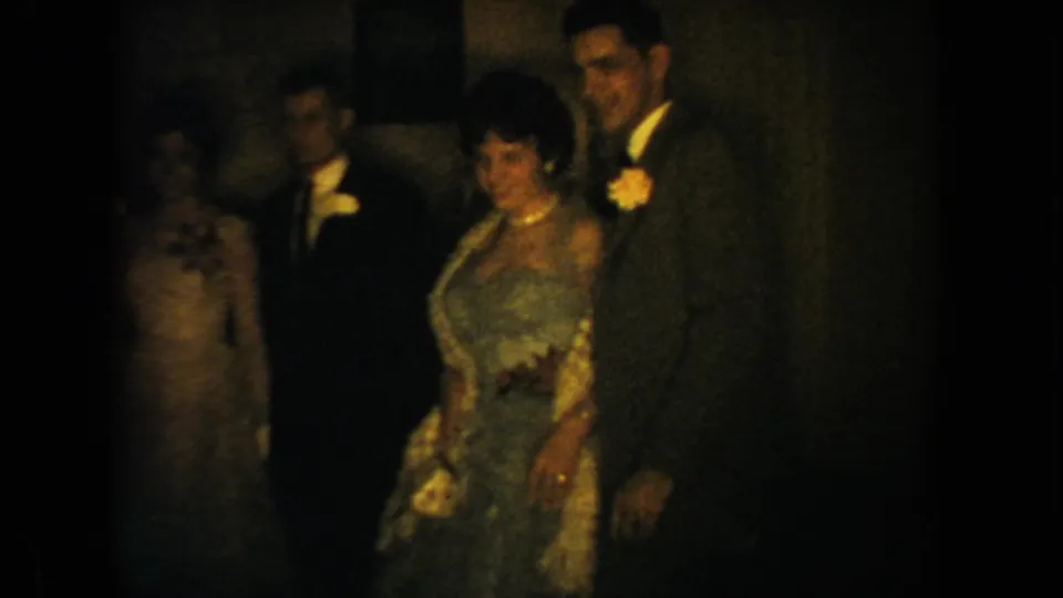A small group of formal guests stands in a dark interior, with a woman in a blue dress in front and suited men nearby.