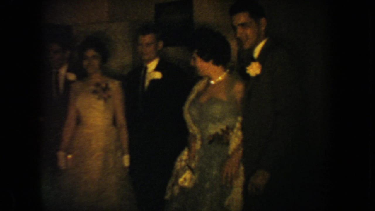 South Dakota 1954 home-movie frame showing wedding reception guests in formal attire standing together in a dim interior.