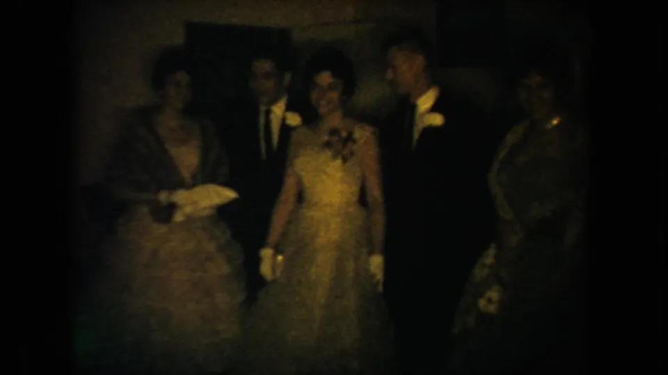 South Dakota 1954 home-movie frame of wedding reception guests gathered close together in low light, some partly obscured by shadow.