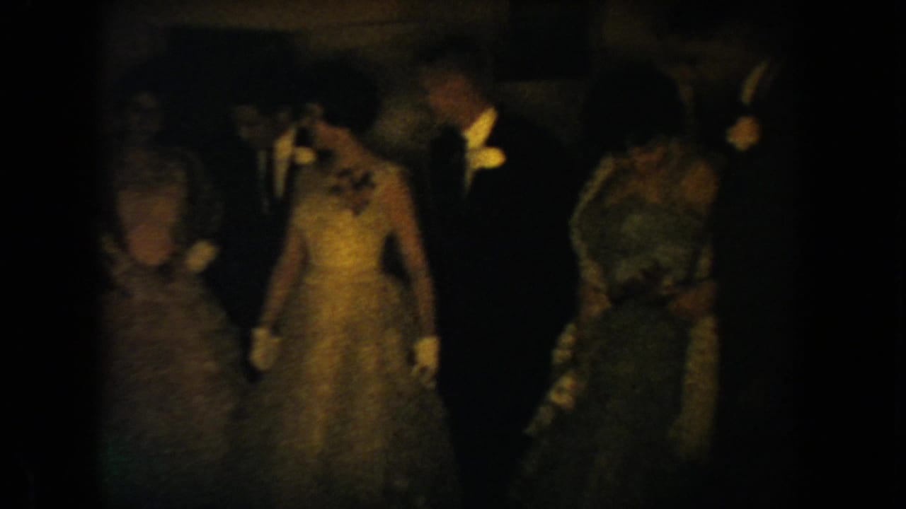 1954 South Dakota home-movie frame showing formally dressed wedding guests in a dark, warm-lit interior.