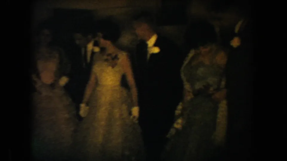 1954 South Dakota home-movie frame showing formally dressed wedding guests in a dark, warm-lit interior.