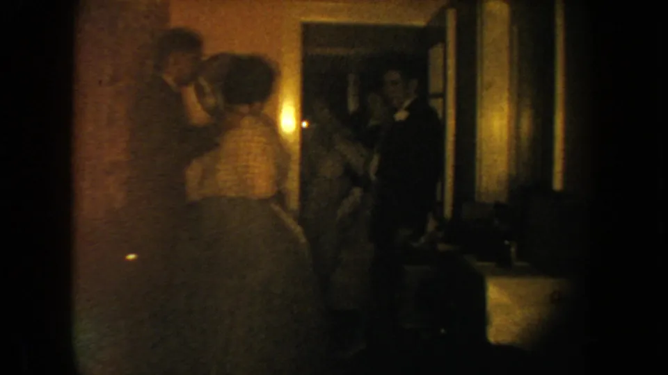 Home-movie frame from a 1954 South Dakota wedding reception showing guests near an interior doorway and wall light.