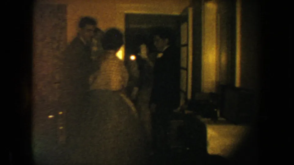 1954 South Dakota wedding reception frame with a foreground couple and guests gathered by a doorway in low light.