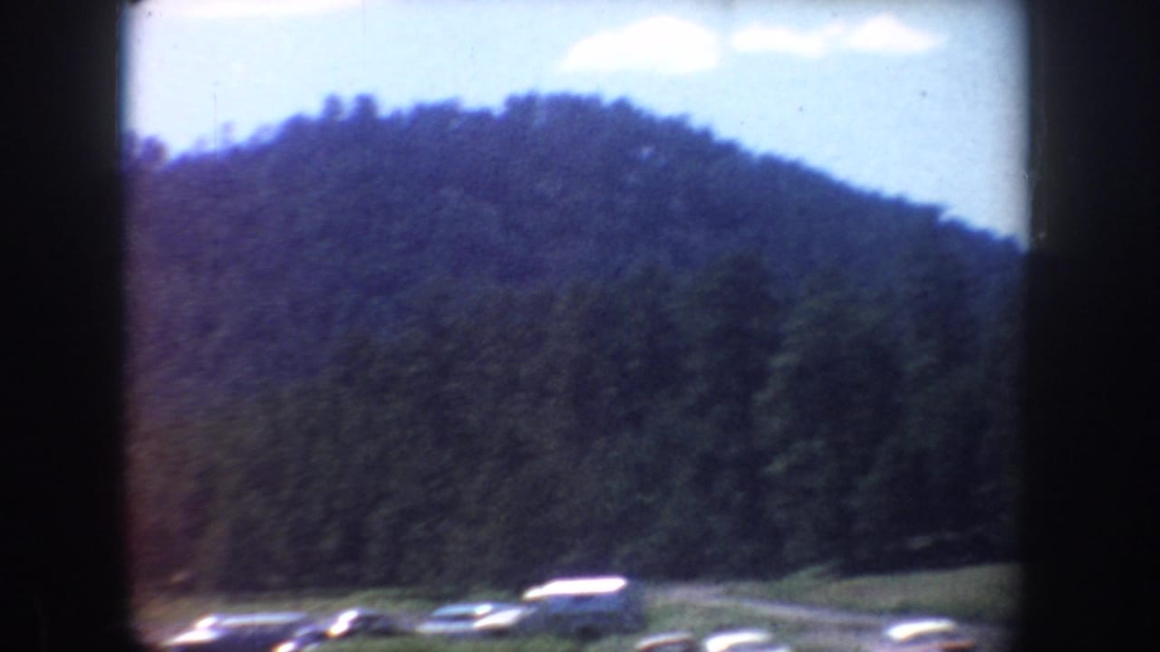 1954 South Dakota home-movie frame showing a wooded hillside and a small roadside parking area with classic cars below.