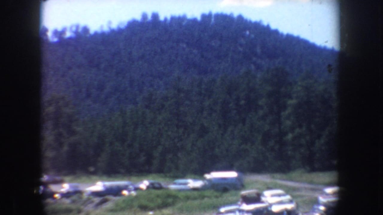 1954 South Dakota frame of a wooded hill above a roadside parking area, with several mid-century cars visible near the treeline.