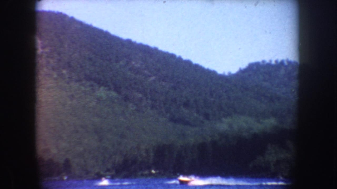 1954 South Dakota home-movie view of a motorboat crossing a lake, backed by dense forest and sloping hills.