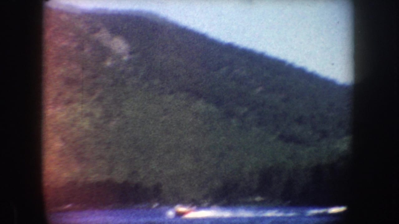 1954 South Dakota frame showing a small motorboat leaving a white wake across a lake beneath a forested slope.