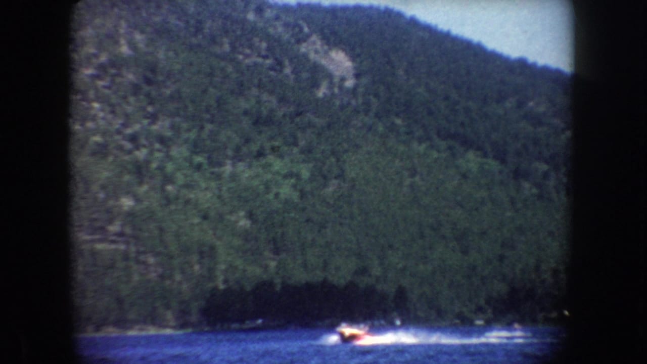 1954 South Dakota home-movie frame showing a distant motorboat leaving a white wake on a lake beneath wooded hills.
