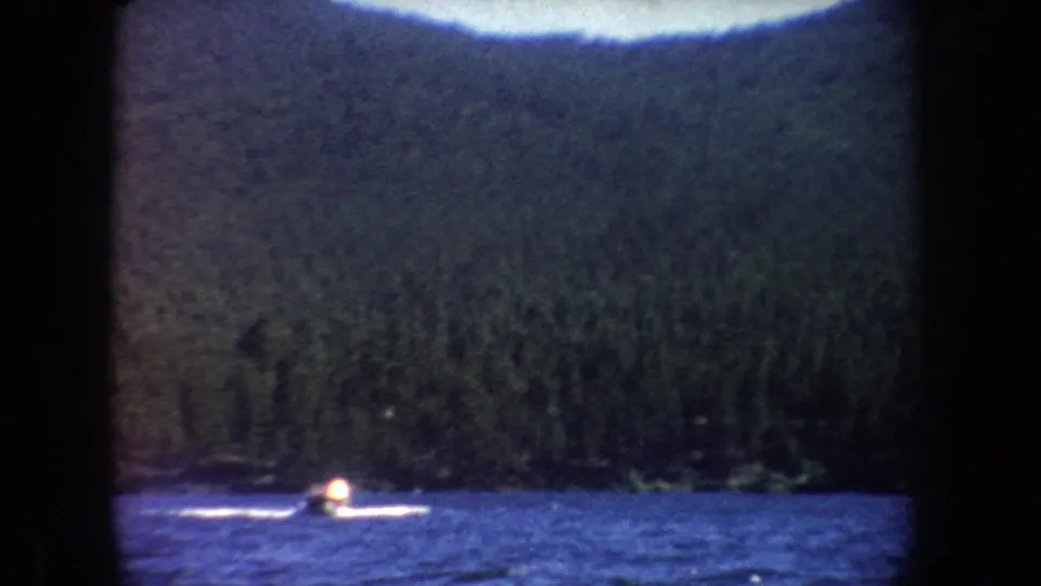 Soft 8mm frame of a small motorboat farther across the lake, with a white wake against dark shoreline trees.