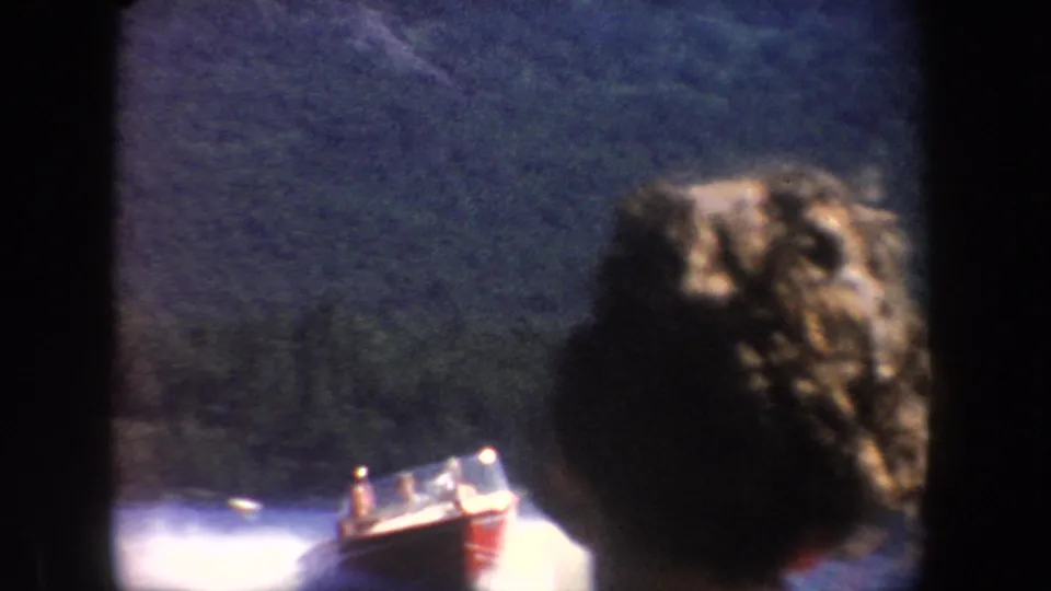 1954 South Dakota 8mm frame showing a red speedboat on a lake, partly obscured by a foreground passenger and dark film borders.