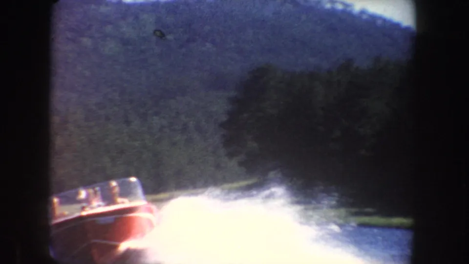 Red speedboat racing across a South Dakota lake in 1954, with bright spray, wooded shoreline, and soft 8mm color.