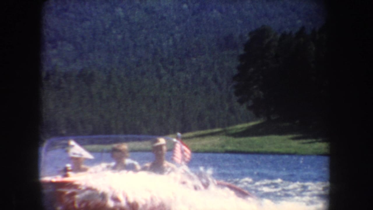 Passengers in a red speedboat ride across a South Dakota lake in 1954, with white spray and a wooded shoreline in the background.