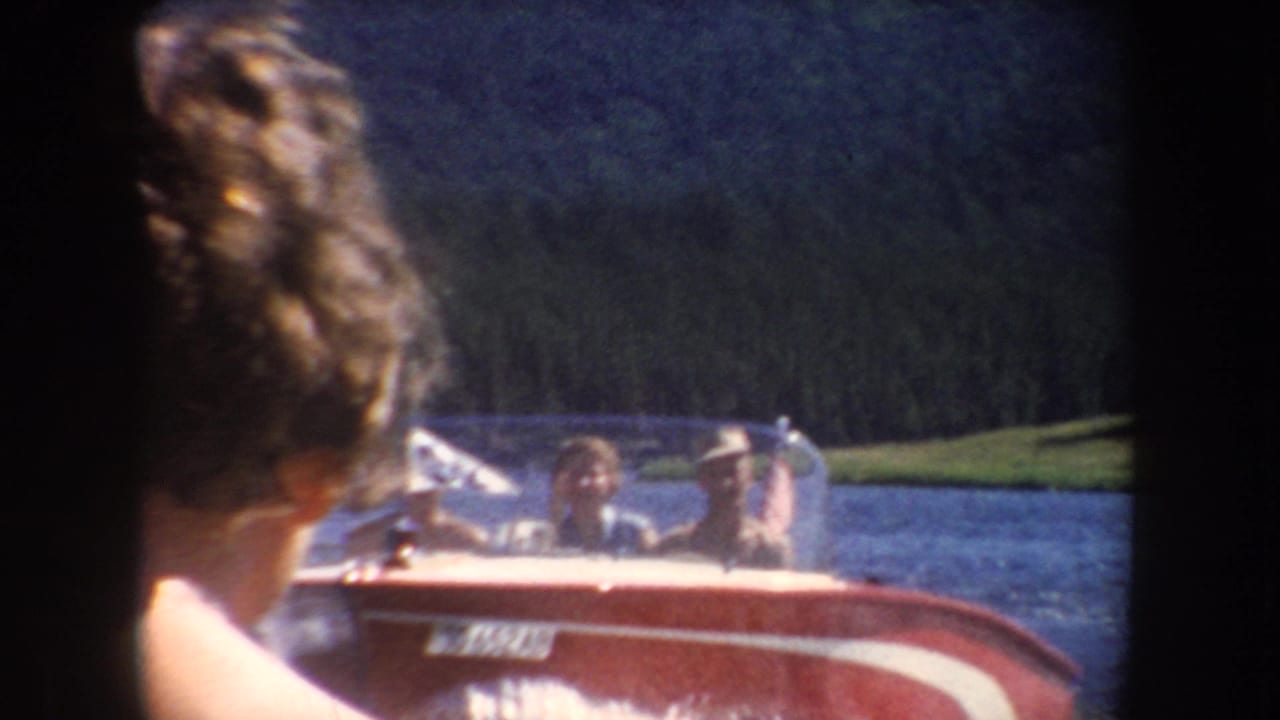 1954 South Dakota home-movie frame showing a red motorboat with passengers moving across a lake, with a woman partly visible in the foreground.
