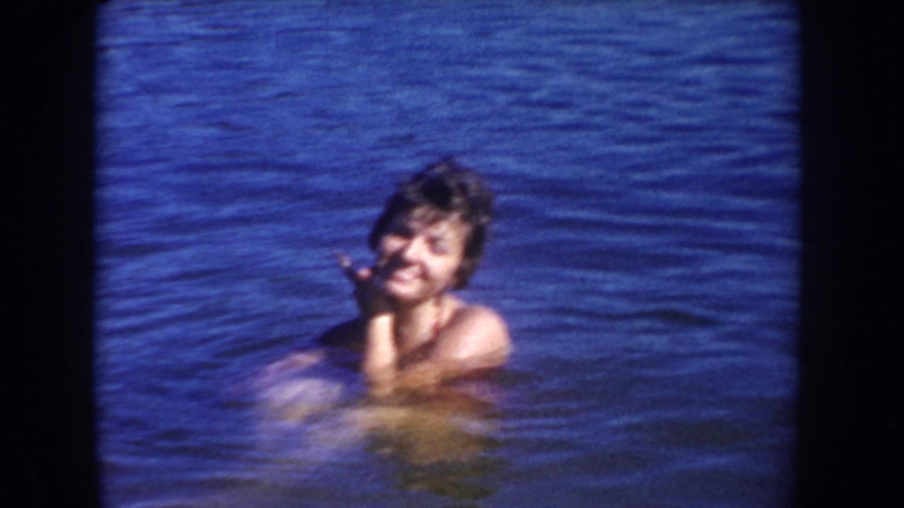 1954 South Dakota home-movie frame of a woman smiling in blue lake water during a sunny swimming scene.