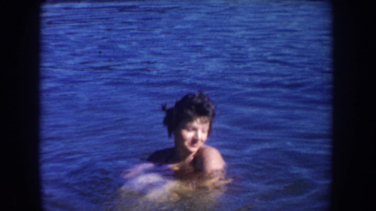 1954 South Dakota home-movie frame showing a woman in the lake looking toward the camera as she swims in bright blue water.