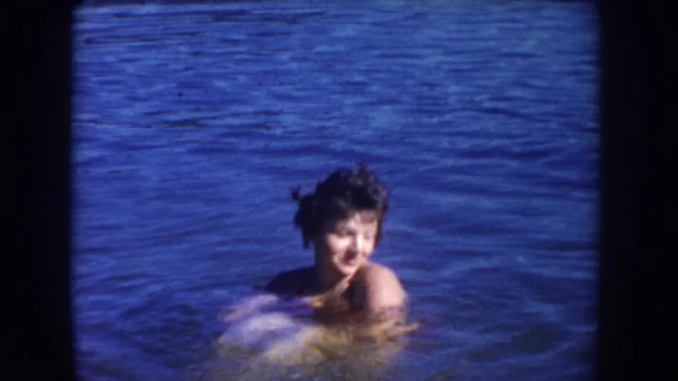 1954 South Dakota home-movie frame showing a woman in the lake looking toward the camera as she swims in bright blue water.