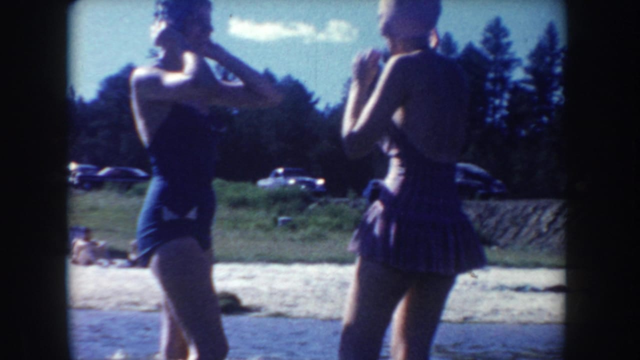 Two young women in 1950s-style swimsuits stand near a South Dakota river while cars sit on a bank behind them.