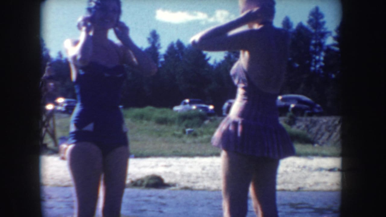 Two women adjust their hair beside a river, with cars and trees in the background.