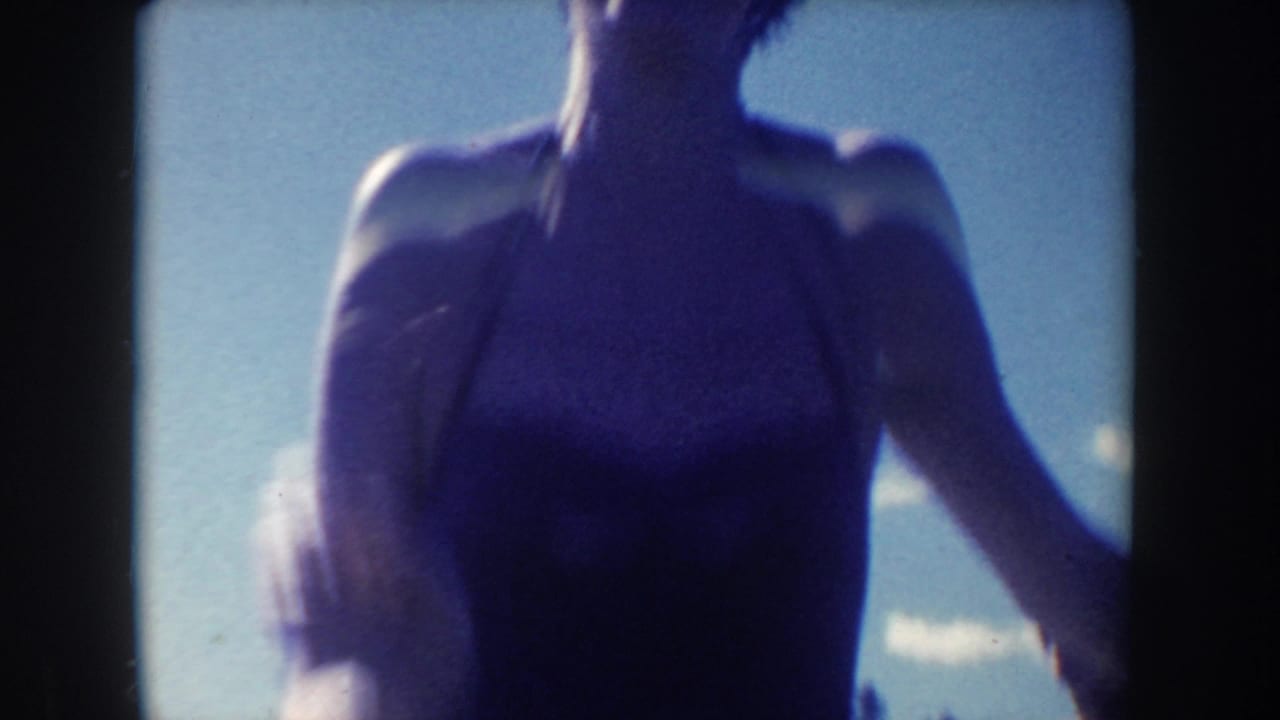 A soft handheld 8mm close view shows a woman in a one-piece swimsuit moving near the river under bright sky.