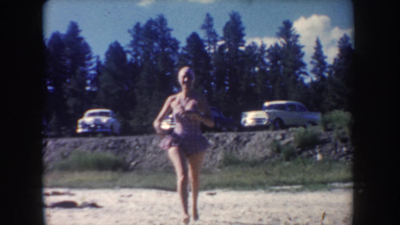 A woman in a mid-century swimsuit moves along the South Dakota riverbank, with parked 1950s cars and trees behind her.