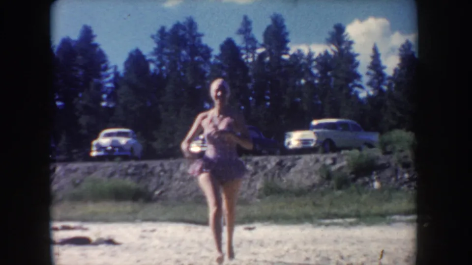 A woman in a mid-century swimsuit moves along the South Dakota riverbank, with parked 1950s cars and trees behind her.