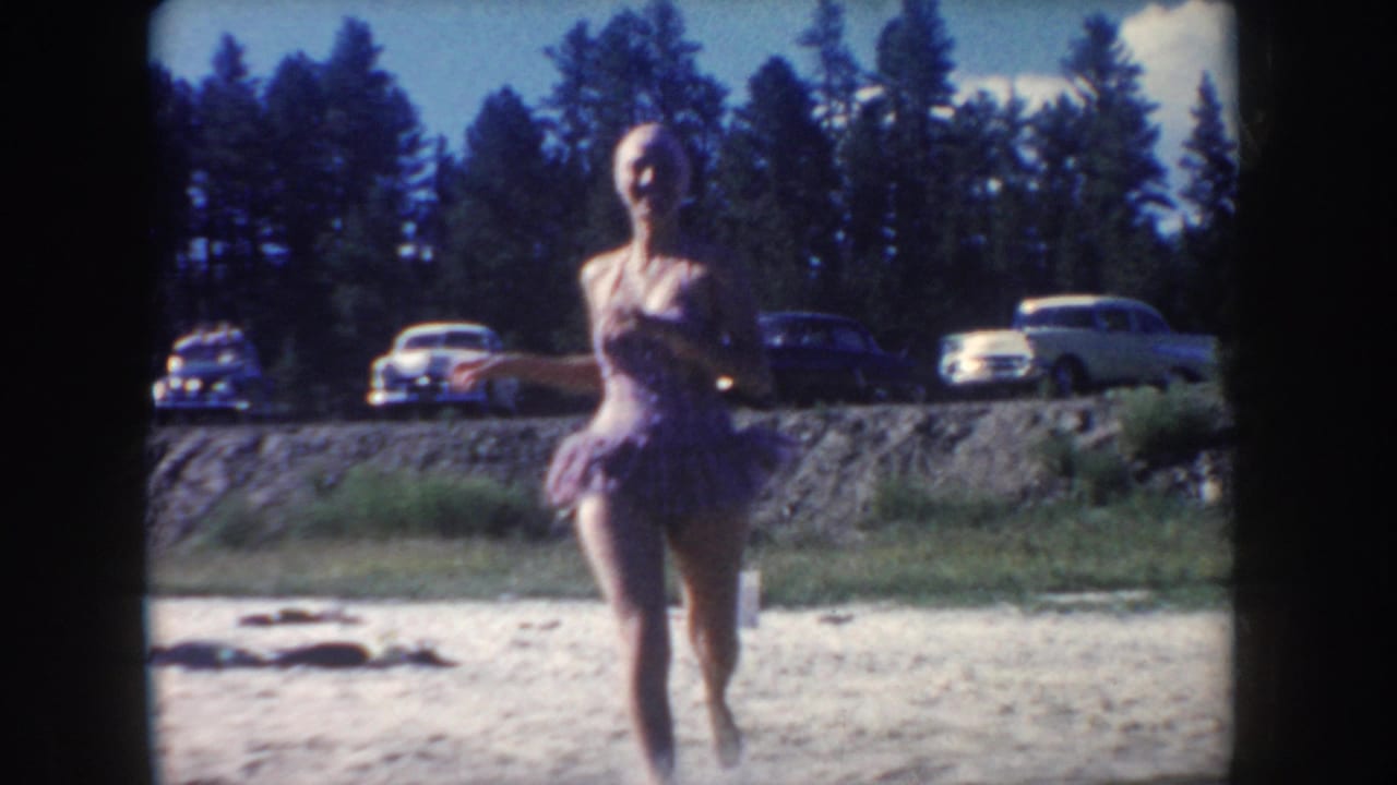 In 1954 South Dakota home-movie footage, a woman runs toward the camera near a lakeside road with parked mid-century cars and pine trees behind her.