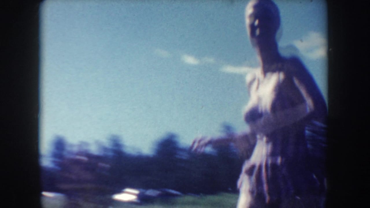 A soft, handheld 8mm frame shows the woman in a purple swimsuit moving close to the camera, with sky and distant trees behind her.