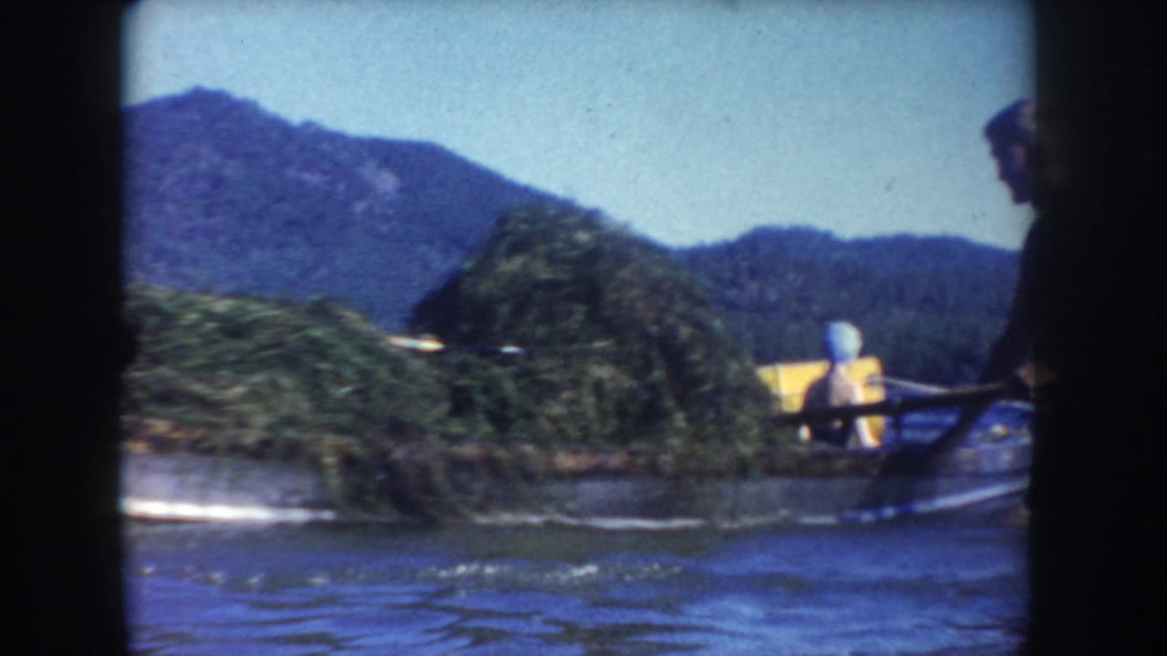 A small boat moves or rests on lake water with adults visible nearby, backed by wooded mountains in a 1950s home-movie frame.