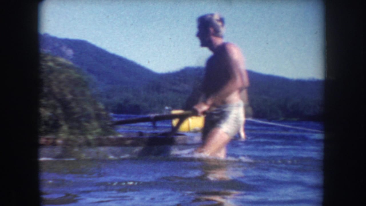 Adult man in swim trunks wades through a South Dakota lake in 1954, with a yellow buoy, wooded shore, and hills behind him.
